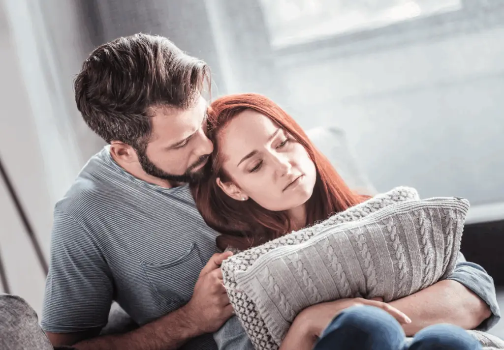 Two people sitting together on a couch with one offering comfort and support on the Trauma Therapy page