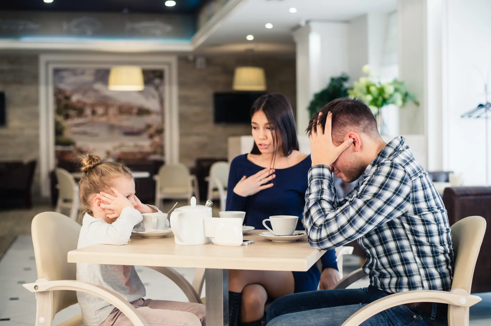 Family sitting at a table with a child covering their face and adults looking tense