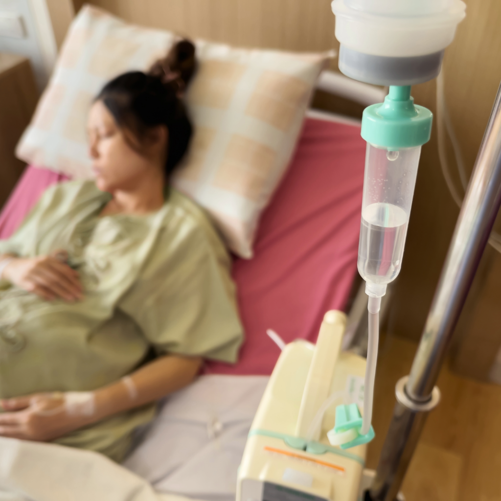 Patient resting in a hospital bed with an IV drip connected to a nearby infusion pump in the foreground