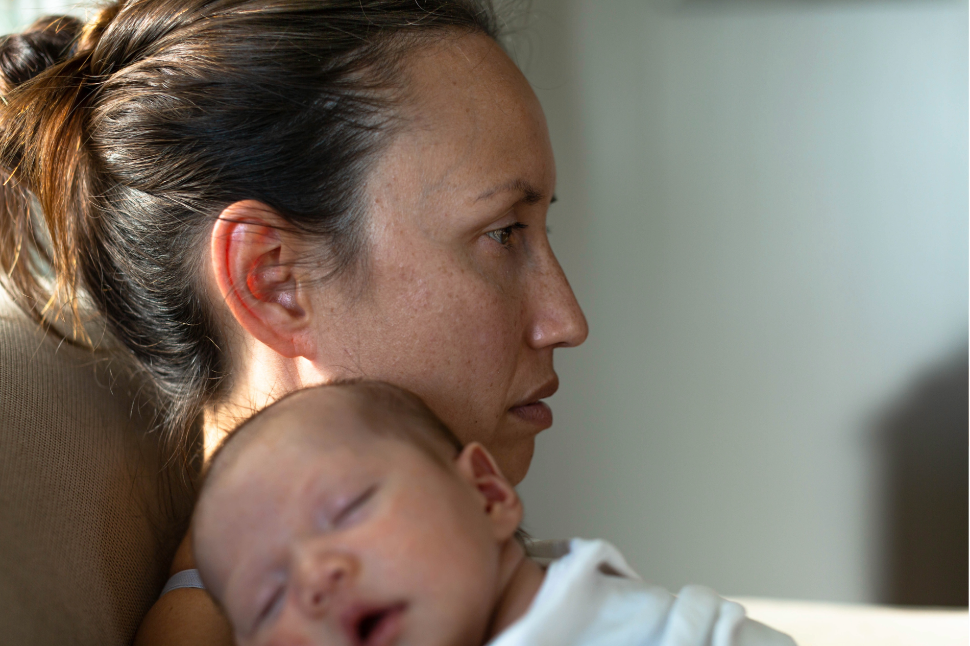 Side profile of a woman holding a sleeping baby close to her chest with calm expression there is soft lighting in the background